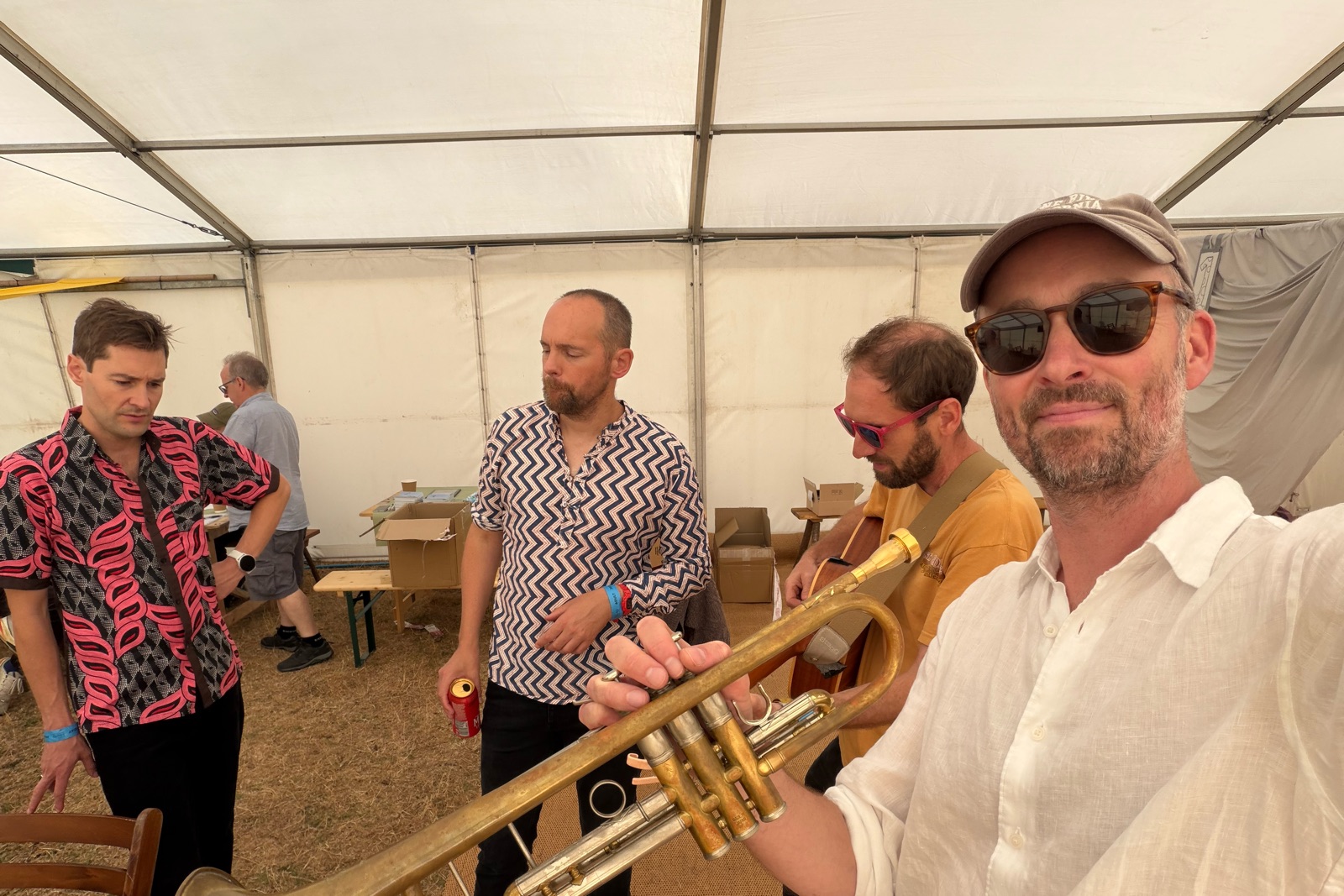 Thomas Hodgson in sunglasses holding a trumpet, with bandmates behind him in a backstage tent.