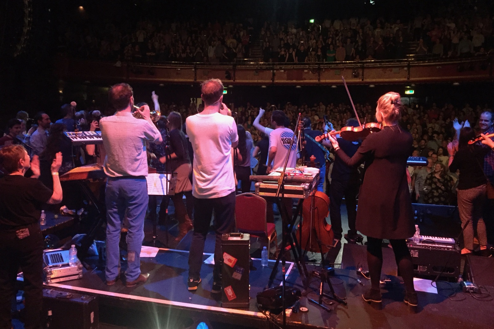 The band from behind on a theatre stage, facing a standing, applauding audience.
