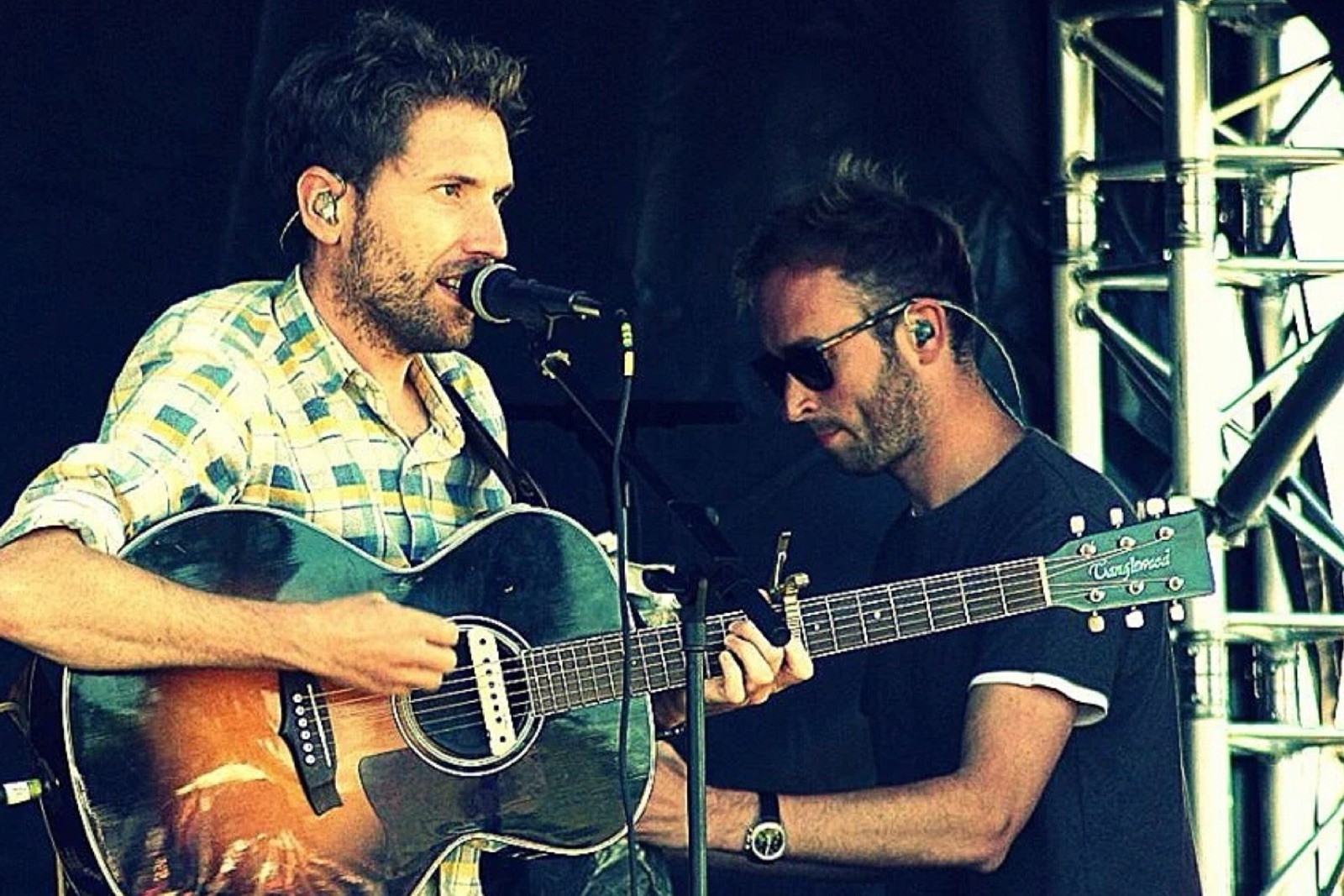 Two members of the band playing acoustic and electric guitar on an outdoor festival stage.