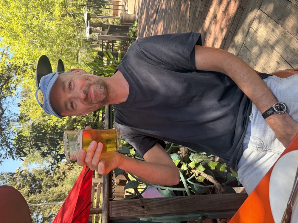 Thomas Hodgson, smiling, raising a glass of beer at an outdoor table under trees.
